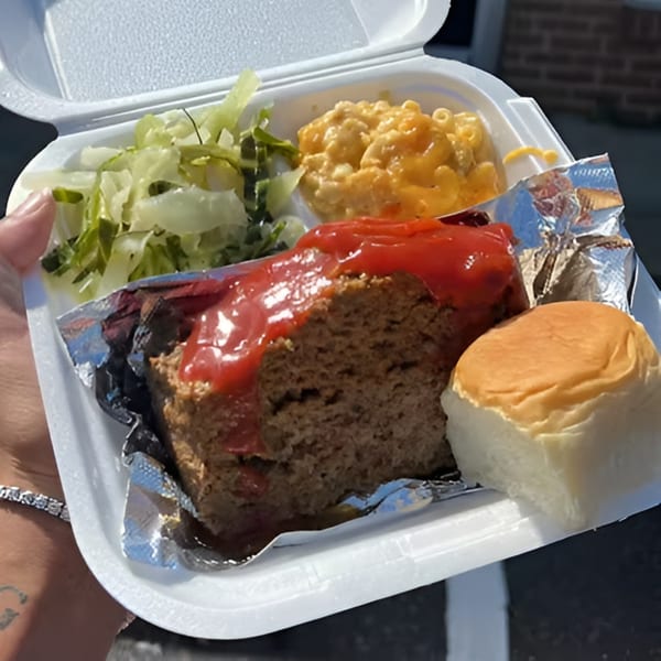 A soul food meatloaf meal with mac and cheese, pickles, and a roll in a to-go container.